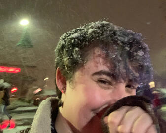 A picture of me at night in a brown coat covered in snow smiling into my coat sleeve
