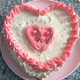 A picture of a white frosted heart shaped cake with a pick heart in the middle and pink details around the edge